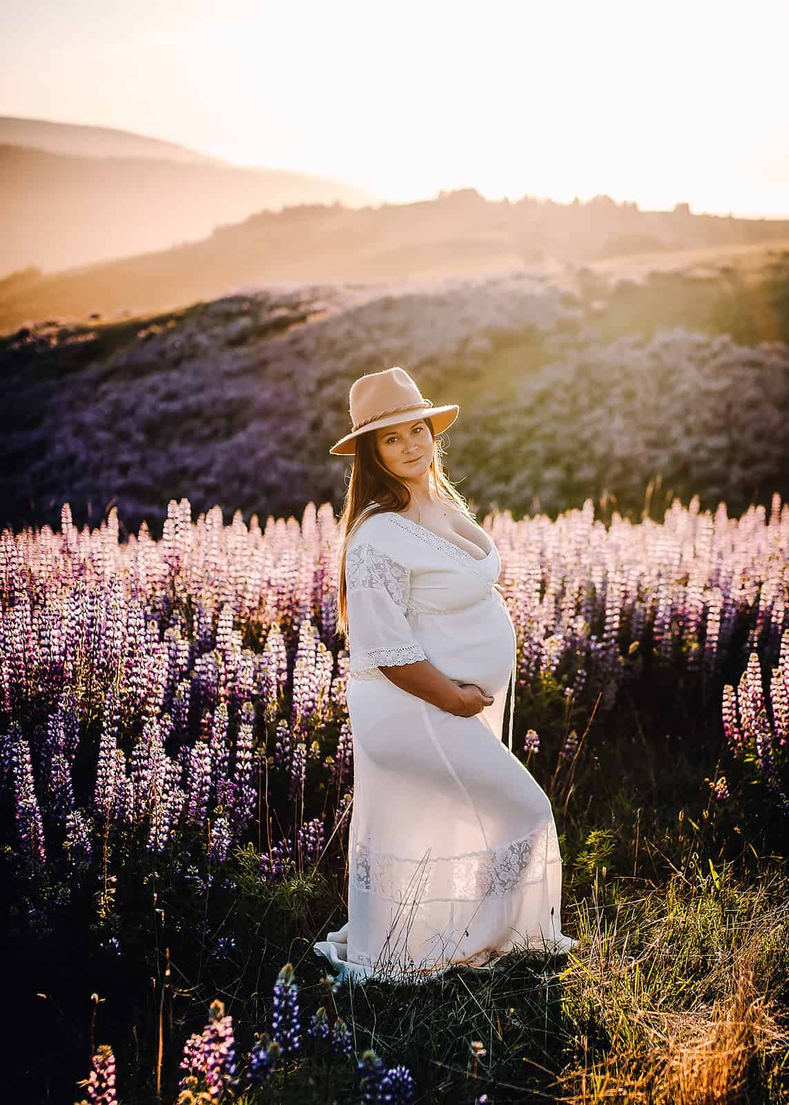 Mom to be in a white gown and hat standing in a lavender field organic elements spa medford