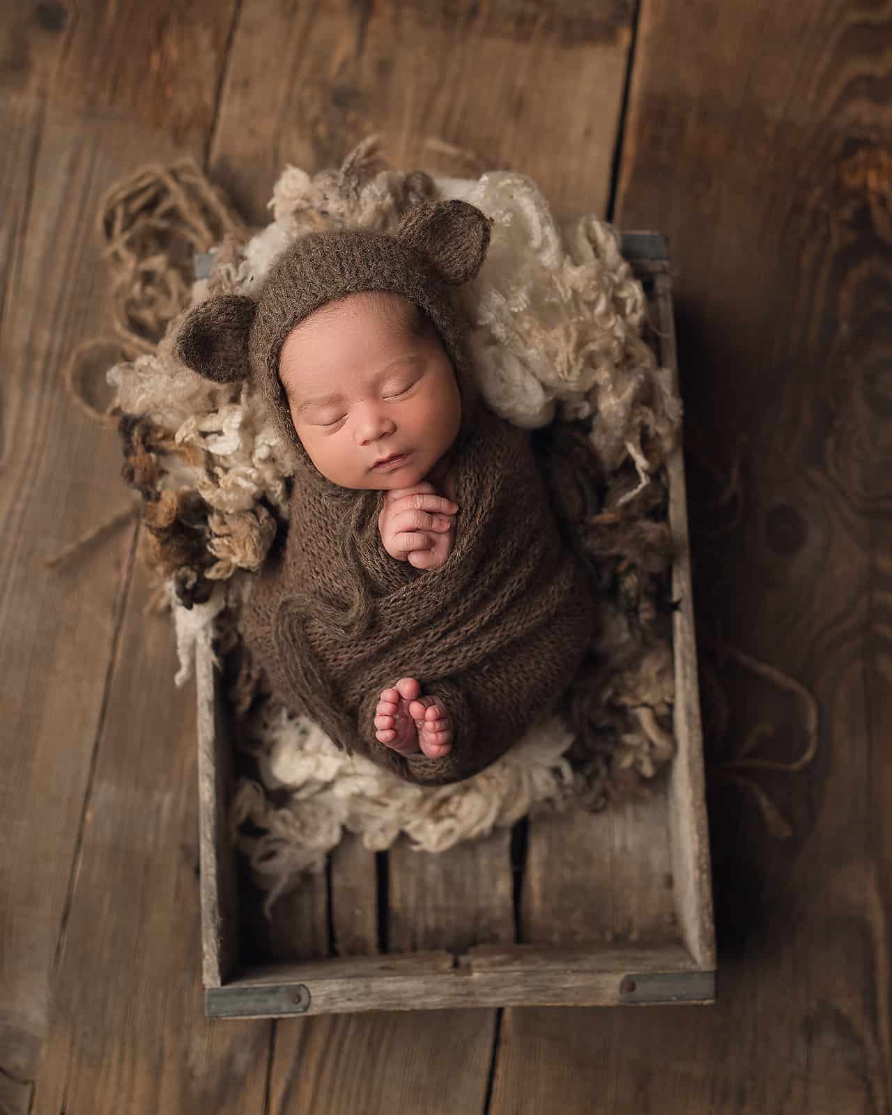 newborn baby in brown wrap and bear ears sleeping in a crate